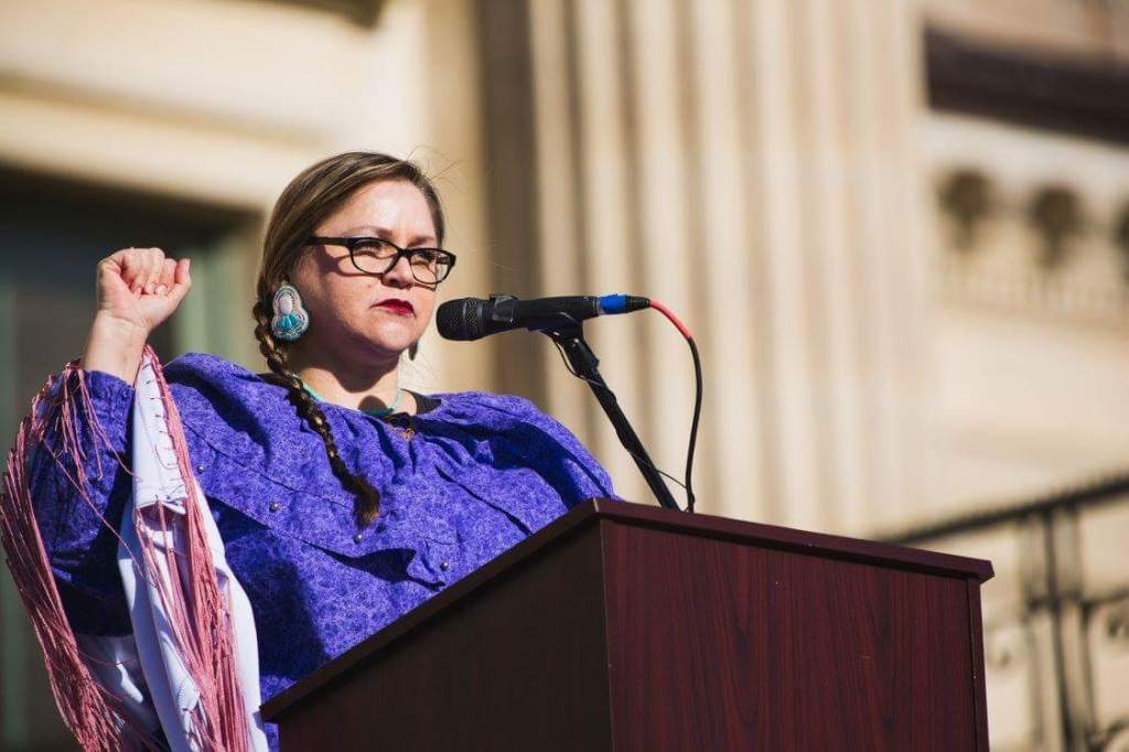 Paulette Blanchard, wearing purple and raising her fist as she speaks from behind a podium. 