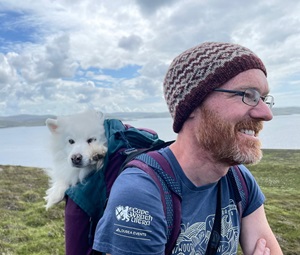 A man with a brown and gray beard and glasses, wearing a blue t-shirt and a beanie, stands outdoors in front of a body of water. A small white dog pokes its head out of the man's backpack. 